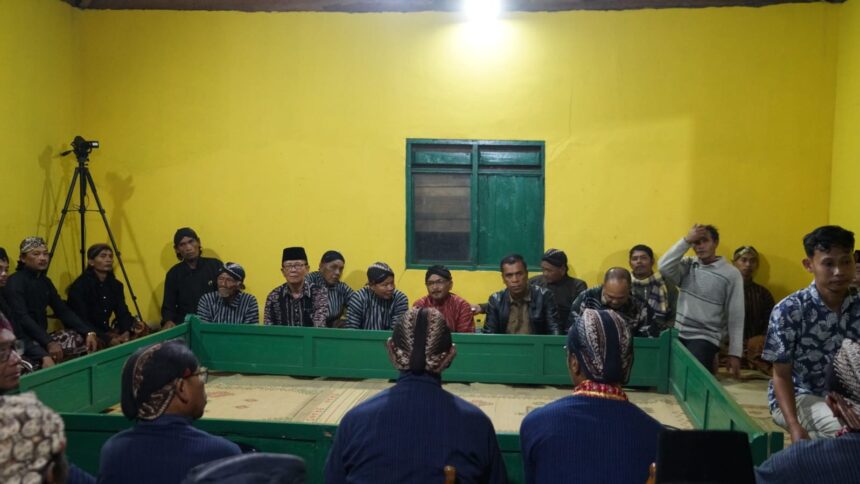 Group of men in traditional attire sit along a green rectangular barrier in a bright yellow room, with a camera on a tripod to the left.
