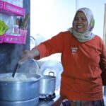 A woman in an orange shirt and hijab stirs a large pot of rice on a kitchen stove, smiling as she cooks.
