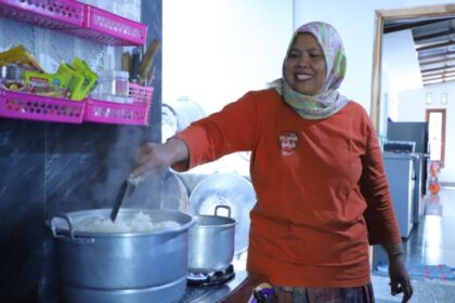 A woman in an orange shirt and hijab stirs a large pot of rice on a kitchen stove, smiling as she cooks.