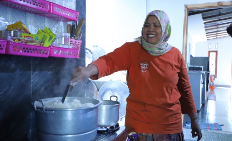 A woman in an orange shirt and hijab stirs a large pot of rice on a kitchen stove, smiling as she cooks.