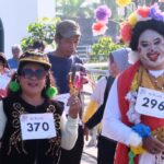 Clown in colorful costume and flower headdress beside parade participants wearing bibs 370 and 296.