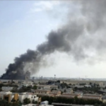 Dark plume of smoke rising from a fire in a distant urban area, over low buildings and a flat landscape.