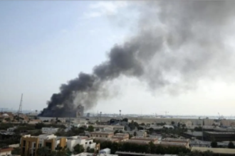 Dark plume of smoke rising from a fire in a distant urban area, over low buildings and a flat landscape.