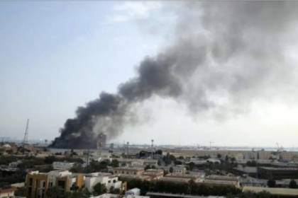 Dark plume of smoke rising from a fire in a distant urban area, over low buildings and a flat landscape.