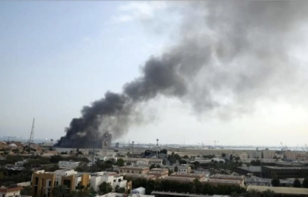Dark plume of smoke rising from a fire in a distant urban area, over low buildings and a flat landscape.