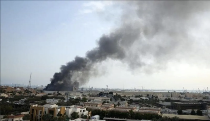 Dark plume of smoke rising from a fire in a distant urban area, over low buildings and a flat landscape.