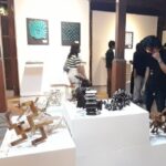 Visitors in an art gallery view white pedestals displaying abstract wooden sculptures, with wall art in the background.