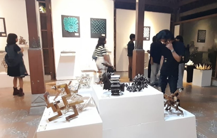 Visitors in an art gallery view white pedestals displaying abstract wooden sculptures, with wall art in the background.