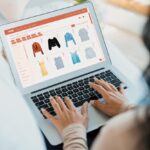 Person typing on a laptop browsing an online clothing store with a grid of dresses and shirts.
