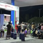Night-time gas station scene with a crowd of people lining up with suitcases and bags in front of fueling stalls.
