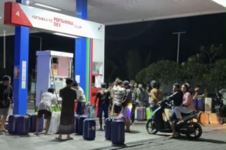 Night-time gas station scene with a crowd of people lining up with suitcases and bags in front of fueling stalls.