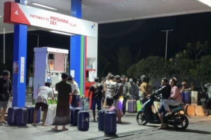 Night-time gas station scene with a crowd of people lining up with suitcases and bags in front of fueling stalls.
