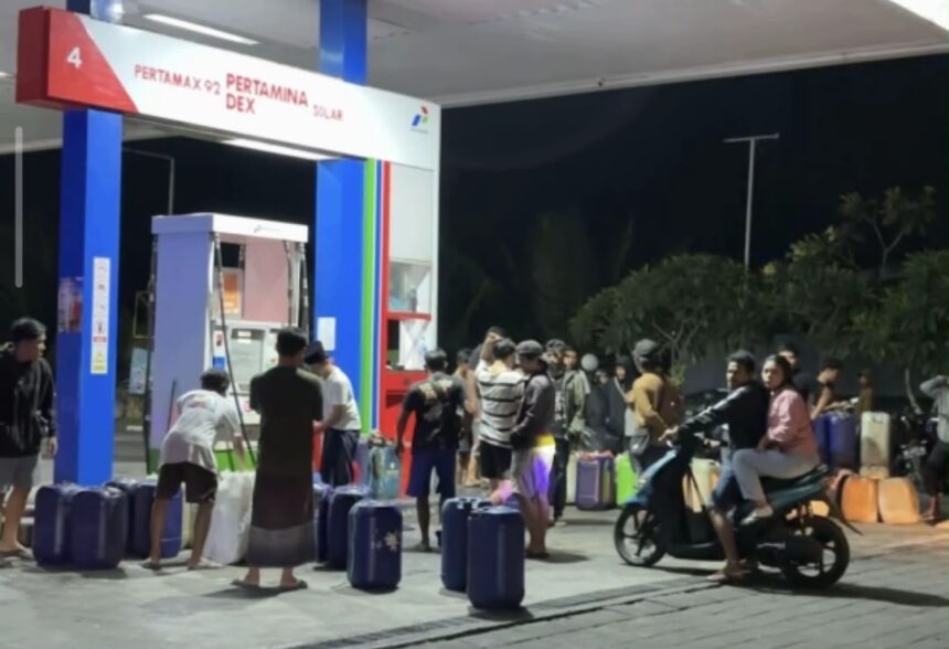 Night-time gas station scene with a crowd of people lining up with suitcases and bags in front of fueling stalls.