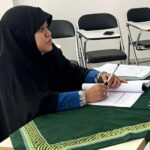 Person in a black hijab and blue sleeves signs papers at a table covered with a green patterned cloth; empty chairs in a room behind.