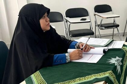 Person in a black hijab and blue sleeves signs papers at a table covered with a green patterned cloth; empty chairs in a room behind.