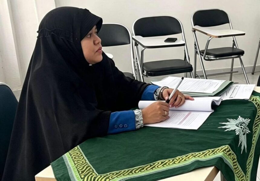 Person in a black hijab and blue sleeves signs papers at a table covered with a green patterned cloth; empty chairs in a room behind.