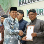 Two men in traditional attire shake hands while receiving certificates at a formal ceremony, with a green banner in the background and others in line holding documents beside them.