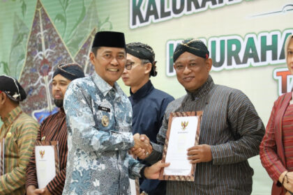 Two men in traditional attire shake hands while receiving certificates at a formal ceremony, with a green banner in the background and others in line holding documents beside them.