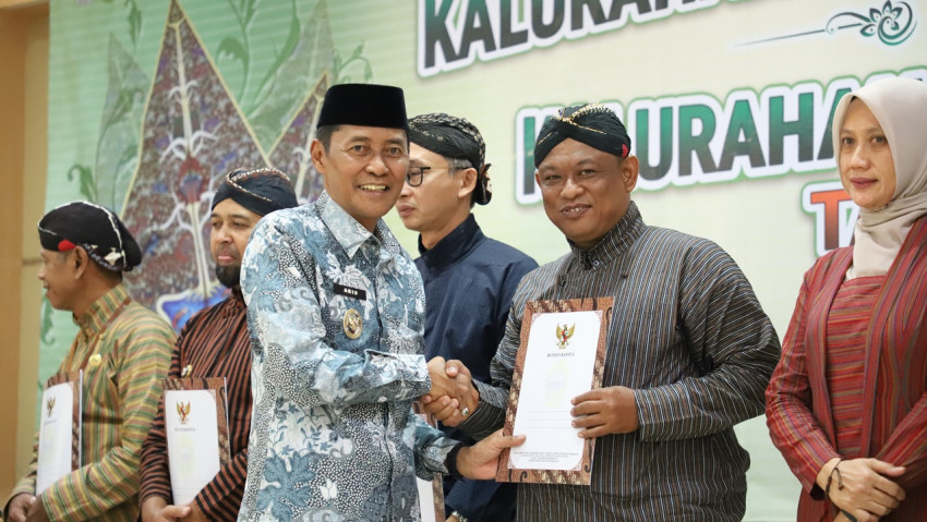 Two men in traditional attire shake hands while receiving certificates at a formal ceremony, with a green banner in the background and others in line holding documents beside them.