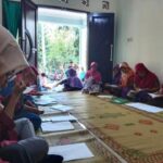 Group of women wearing colorful headscarves sit on a carpeted floor, writing in notebooks during a community class session.