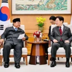 Two officials in formal suits sit in armchairs during a diplomatic meeting, with flags behind them and a flower arrangement on a small table between them.