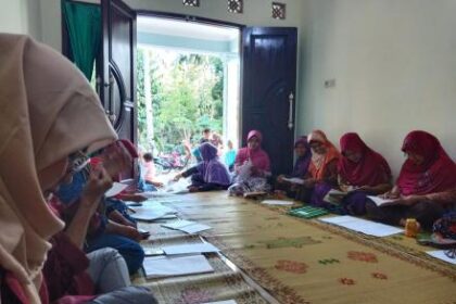 Group of women wearing colorful headscarves sit on a carpeted floor, writing in notebooks during a community class session.
