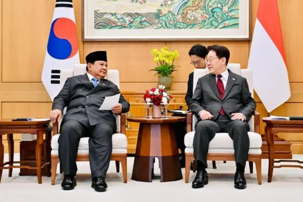 Two officials in formal suits sit in armchairs during a diplomatic meeting, with flags behind them and a flower arrangement on a small table between them.