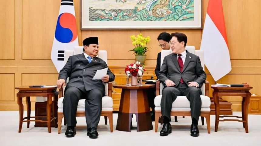 Two officials in formal suits sit in armchairs during a diplomatic meeting, with flags behind them and a flower arrangement on a small table between them.
