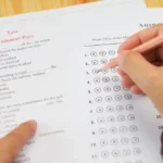 Close-up of a hand filling in bubbles on a multiple-choice answer sheet during a grammar test on a desk with a pink pencil nearby.