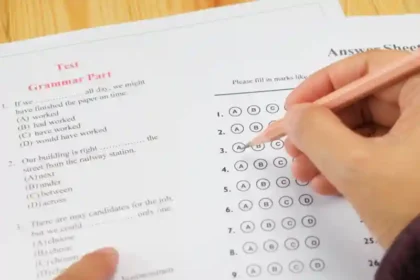 Close-up of a hand filling in bubbles on a multiple-choice answer sheet during a grammar test on a desk with a pink pencil nearby.