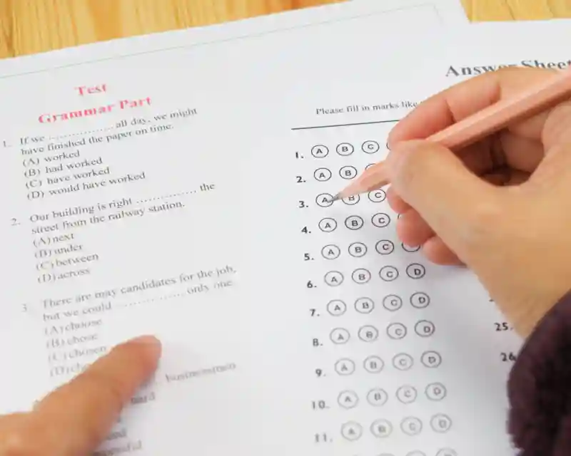 Close-up of a hand filling in bubbles on a multiple-choice answer sheet during a grammar test on a desk with a pink pencil nearby.