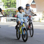 Family of three riding bicycles on a sunny street, with a child on a yellow bike in the center, all wearing helmets.