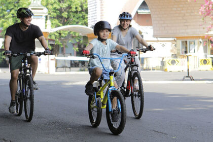 Family of three riding bicycles on a sunny street, with a child on a yellow bike in the center, all wearing helmets.