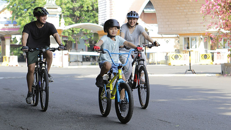 Family of three riding bicycles on a sunny street, with a child on a yellow bike in the center, all wearing helmets.