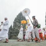Group of Balinese people in white and colorful sarongs walk in a temple procession beside ancient stone towers.