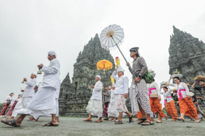 Group of Balinese people in white and colorful sarongs walk in a temple procession beside ancient stone towers.