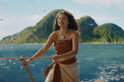 Woman on a boat smiling while holding a rope, with blue sea and a green island in the distance behind her.