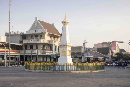 A white ornate monument with gold accents stands inside a circular plaza, fenced by a green barrier, with buildings and street activity in the background.