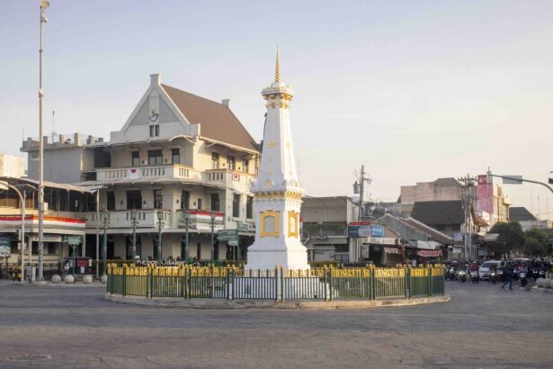 A white ornate monument with gold accents stands inside a circular plaza, fenced by a green barrier, with buildings and street activity in the background.