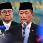 Man in a dark suit and blue tie speaks into a microphone at a press conference, with reporters' microphones in front and another man in the background wearing a traditional cap.
