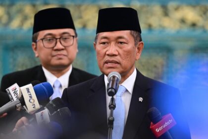 Man in a dark suit and blue tie speaks into a microphone at a press conference, with reporters' microphones in front and another man in the background wearing a traditional cap.