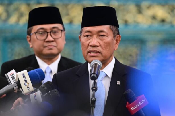 Man in a dark suit and blue tie speaks into a microphone at a press conference, with reporters' microphones in front and another man in the background wearing a traditional cap.