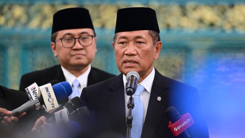 Man in a dark suit and blue tie speaks into a microphone at a press conference, with reporters' microphones in front and another man in the background wearing a traditional cap.