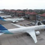 Garuda Indonesia airplane at a gate on a busy airport apron, with other parked planes and a terminal building in the background.