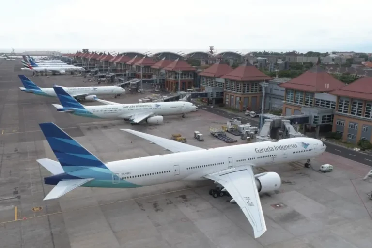 Garuda Indonesia airplane at a gate on a busy airport apron, with other parked planes and a terminal building in the background.