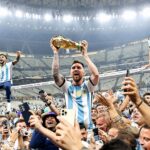 Soccer players celebrate a championship, lifting a golden trophy above a cheering crowd in a stadium.