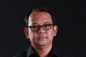 Head-and-shoulders portrait of a man wearing a black shirt and glasses against a dark background.