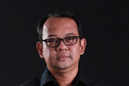 Head-and-shoulders portrait of a man wearing a black shirt and glasses against a dark background.