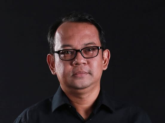 Head-and-shoulders portrait of a man wearing a black shirt and glasses against a dark background.