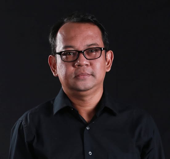 Head-and-shoulders portrait of a man wearing a black shirt and glasses against a dark background.